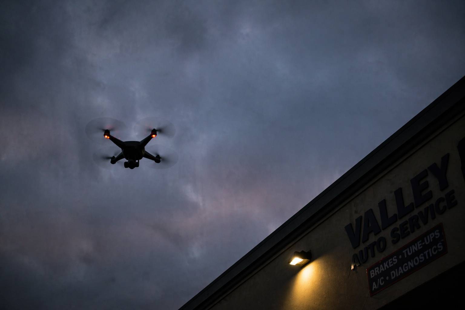Drone silhouetted against a cloudy dusk sky hovering above a commercial building, viewed from a low angle in an urban business area